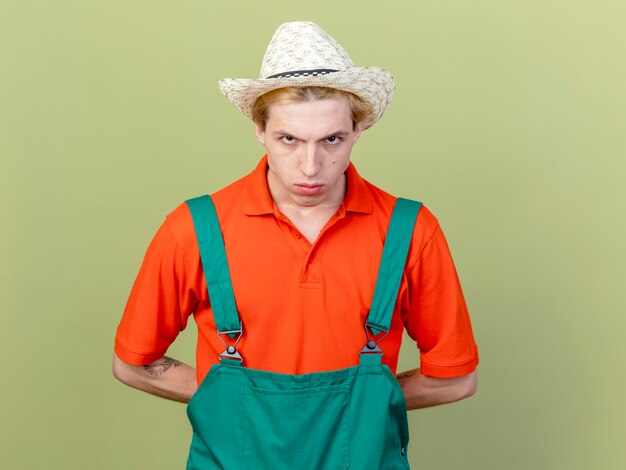 young gardener man wearing jumpsuit and hat with serious face