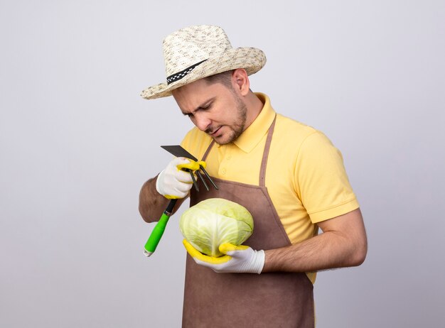 Young gardener man wearing jumpsuit and hat in working gloves holding cabbage and mattock looking with serious face 