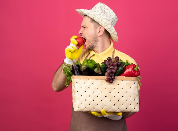 新鮮なトマトを噛む野菜でいっぱいの木枠を保持している作業用手袋でジャンプスーツと帽子を身に着けている若い庭師の男