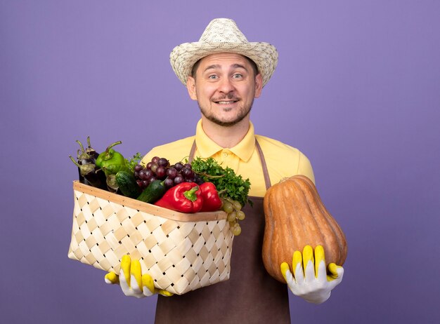 Молодой садовник в комбинезоне и шляпе в рабочих перчатках держит ящик, полный овощей с тыквой, улыбается со счастливым лицом