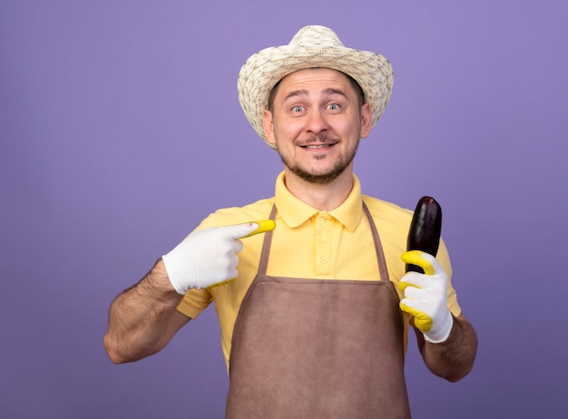 Young gardener man wearing jumpsuit and hat in working gloves holding eggplant pointing with index figner at it smiling with happy face 