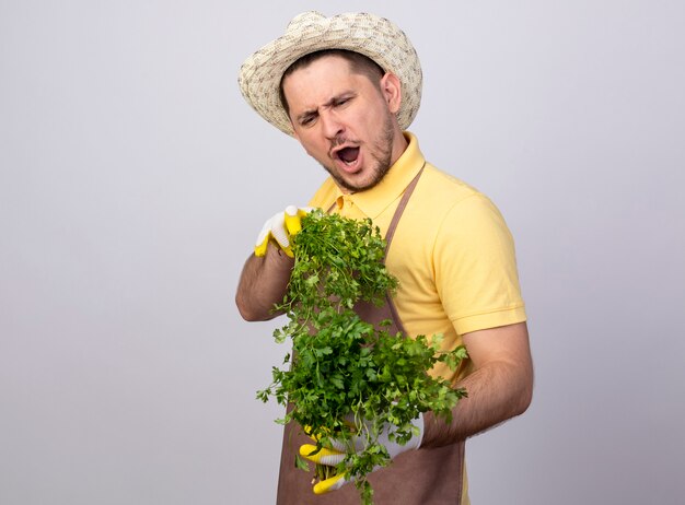 Young gardener man wearing jumpsuit and hat in working gloves holding fresh herbs happy and excited 