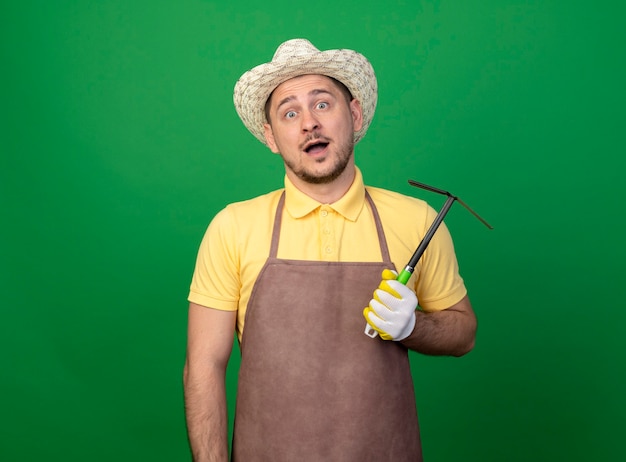 Young gardener man wearing jumpsuit and hat in working gloves holding mattock confused 