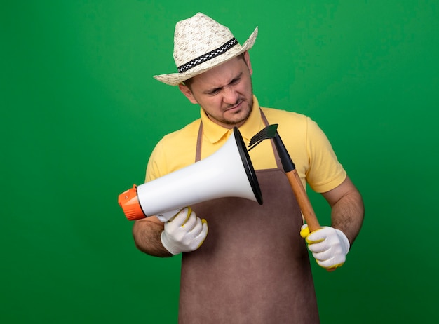 Young gardener man wearing jumpsuit and hat in working gloves holding megaphone scratching it with mini rake 