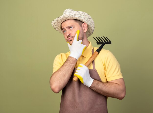 Young gardener man wearing jumpsuit and hat in working gloves holding mini rake looking aside with hand on chin thinking
