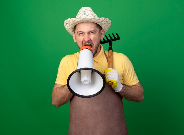 Молодой садовник в комбинезоне и шляпе в рабочих перчатках держит мини-грабли и кричит в мегафон с агрессивным выражением лица