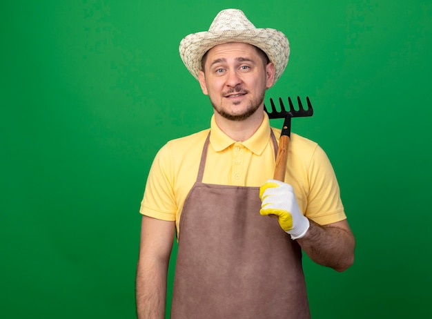 Young gardener man wearing jumpsuit and hat in working gloves holding mini rake smiling cheerfully 
