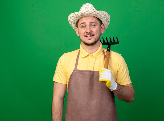 陽気な笑顔のミニ熊手を保持している作業用手袋でジャンプスーツと帽子を身に着けている若い庭師の男