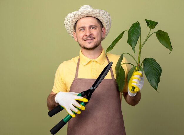 明るい壁の上に立っている顔に笑顔で正面を見て植物と生け垣クリッパーを保持している作業手袋でジャンプスーツと帽子を身に着けている若い庭師の男