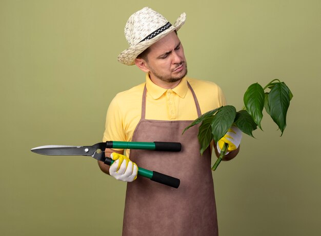 真面目な顔で見ている植物と生け垣クリッパーを保持している作業用手袋でジャンプスーツと帽子を身に着けている若い庭師の男