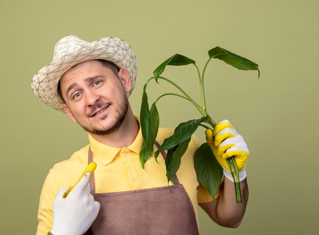 Young gardener man wearing jumpsuit and hat in working gloves holding plant looking at front smiling with happy face standing over light wall
