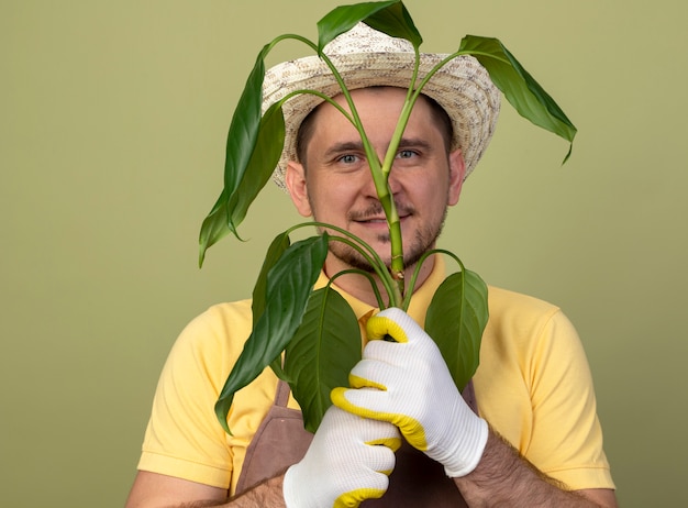 明るい壁の上に立っている幸せそうな顔で笑顔で正面を見て植物を保持している作業用手袋でジャンプスーツと帽子を身に着けている若い庭師の男