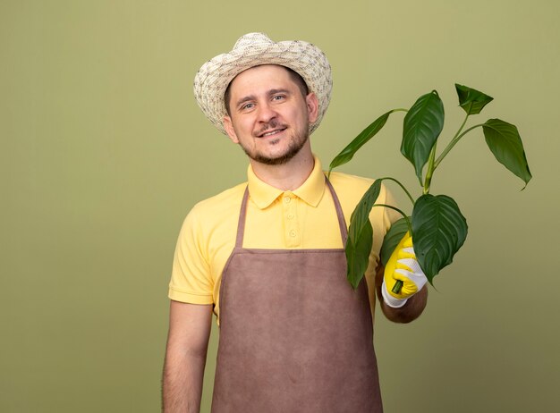 Young gardener man wearing jumpsuit and hat in working gloves holding plant with smile on face 