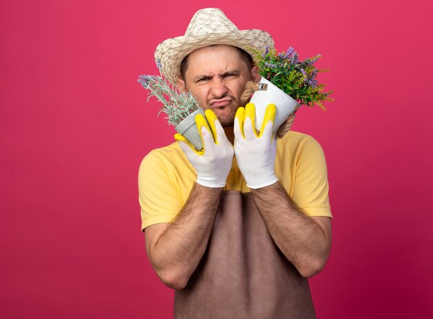 Young gardener man wearing jumpsuit and hat in working gloves holding potted plants being confused and displeased 