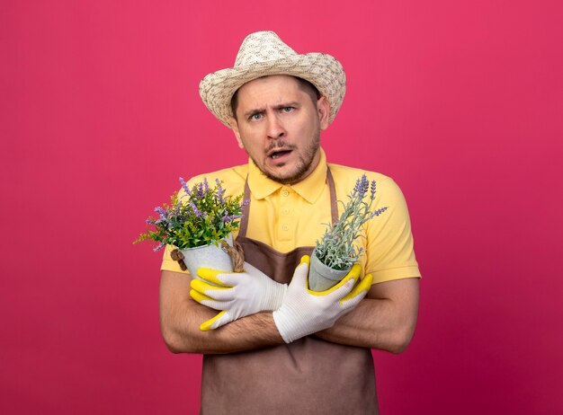 Young gardener man wearing jumpsuit and hat in working gloves holding potted plants confused 