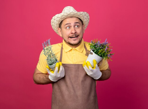混乱している脇を見ている鉢植えの植物を保持している作業用手袋でジャンプスーツと帽子を身に着けている若い庭師の男