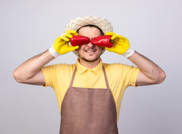 Free photo young gardener man wearing jumpsuit and hat in working gloves holding red bell peppers over his eyes smiling