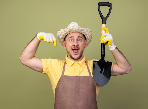 Молодой садовник в комбинезоне и шляпе в рабочих перчатках держит лопату, указывая вниз, весело улыбаясь
