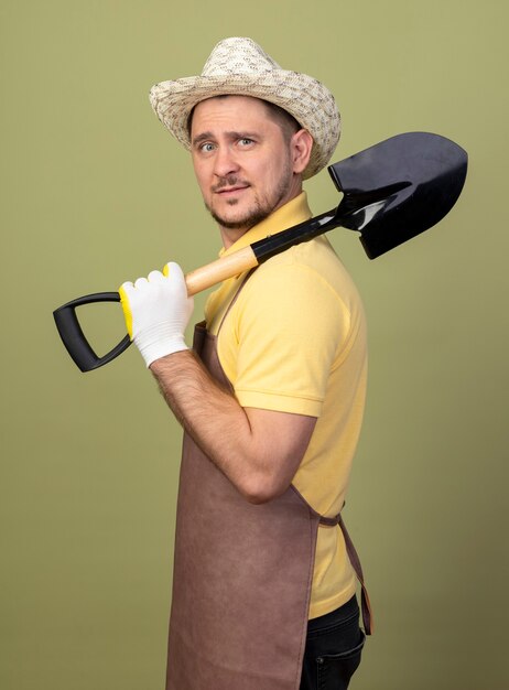 Young gardener man wearing jumpsuit and hat in working gloves holding shovel on shoulder looking confident 