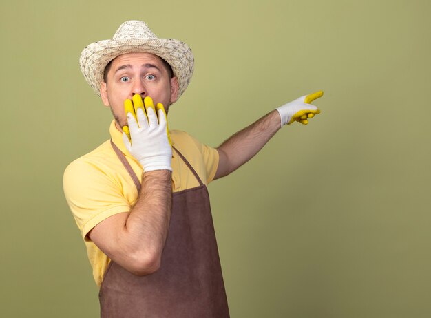 Young gardener man wearing jumpsuit and hat in working gloves pointing with index finger at something being shocked covering mouth with hand 