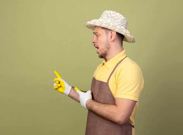 Young gardener man wearing jumpsuit and hat in working gloves pointing with index fingers back looking confused 