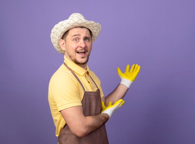 作業用手袋でジャンプスーツと帽子を身に着けている若い庭師の男は、元気に笑顔の腕で何かを提示します。