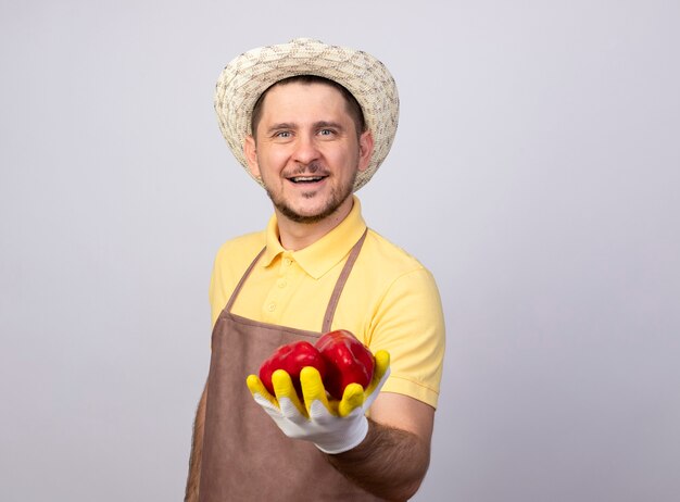 幸せそうな顔で笑顔の赤ピーマンを示す作業手袋でジャンプスーツと帽子を身に着けている若い庭師の男