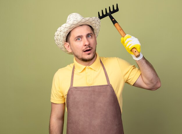 Young gardener man wearing jumpsuit and hat in working gloves swinging mini rake looking at front confused standing over light wall