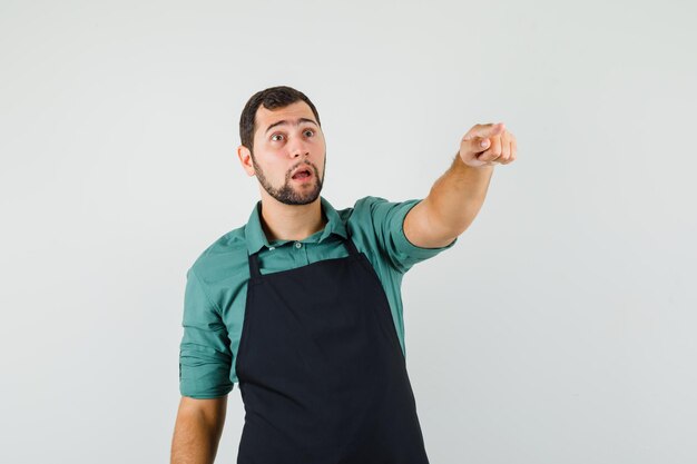 Young gardener pointing away in t-shirt,apron and looking surprised. front view.