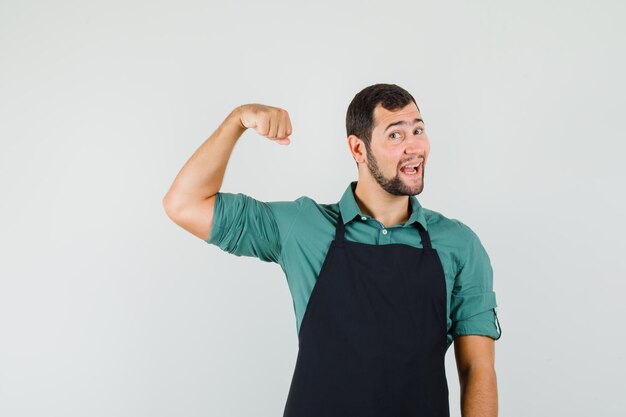 Young gardener showing his arm muscles in t-shirt,apron and looking jolly , front view.
