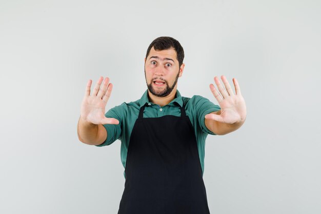 Young gardener showing stop gesture in t-shirt,apron and looking scared. front view.