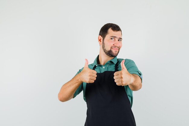 Молодой садовник показывает палец вверх в футболке, фартуке и рад, вид спереди.