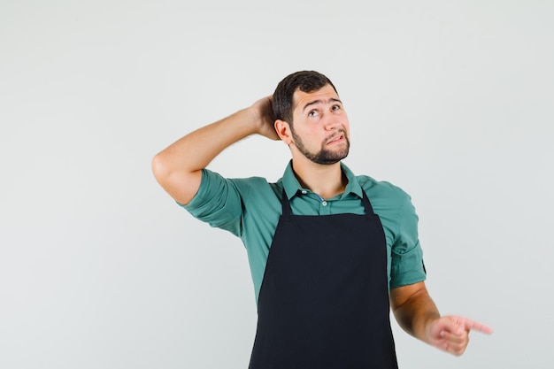 Young gardener in t-shirt,apron holding hand on his head while pointing aside and looking puzzled , front view.