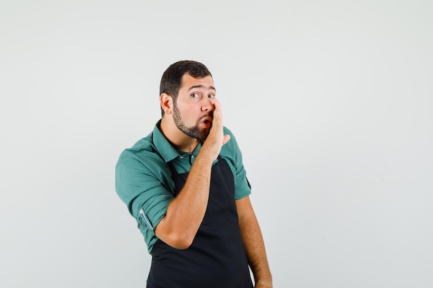 Young gardener in t-shirt,apron saying something secretly and looking focused , front view.