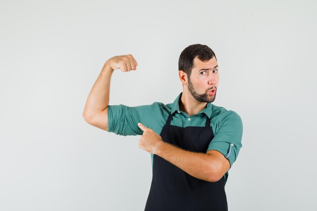 Young gardener in t-shirt,apron showing his arm muscles and looking amazed , front view.