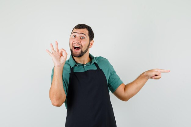 Young gardener in t-shirt,apron showing ok gesture while pointing aside and looking merry , front view.