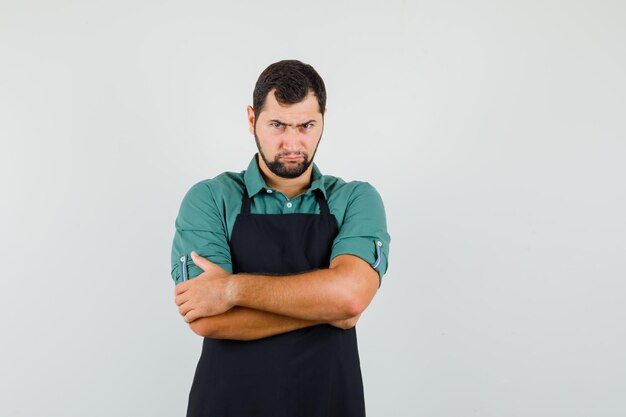 Young gardener in t-shirt,apron standing with crossed arms and looking offended , front view.