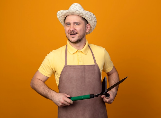 Young gardener wearing jumpsuit and hat holding hedge clippers looking at front smiling with happy face standing over orange wall