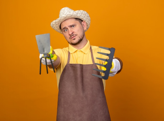 Free photo young gardener wearing jumpsuit and hat holding mattock and mini rake pointing with them at camera looking confident standing over orange wall