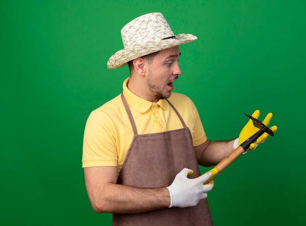 Free photo young gardener wearing jumpsuit and hat holding mini rake looking at it being surprised standing over green wall