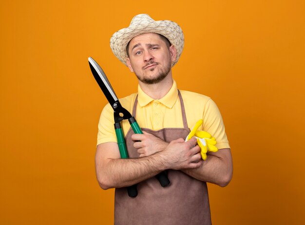 Young gardener wearing jumpsuit and hat holding working gloves and hedge clippers looking at front with confident expression standing over orange wall