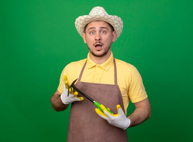 Young gardener wearing jumpsuit and hat in working gloves demonstrating mattock looking at front surprised standing over green wall