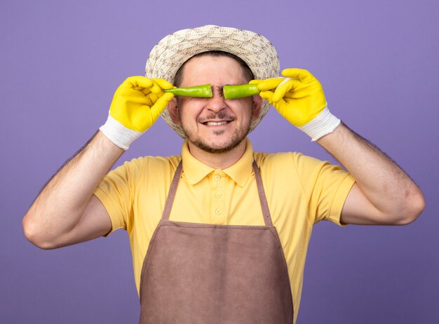 紫色の壁の上に立って笑顔の目の上に壊れた緑の唐辛子を保持している作業手袋でジャンプスーツと帽子を身に着けている若い庭師