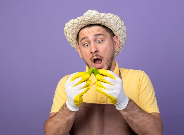 Young gardener wearing jumpsuit and hat in working gloves holding broken green chili pepper looking at it surpried and amazed standing over purple wall