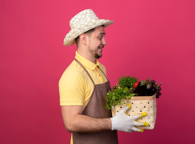 ピンクの壁の上に立って幸せで前向きに野菜を見ている野菜でいっぱいの木枠を保持している作業用手袋でジャンプスーツと帽子を身に着けている若い庭師