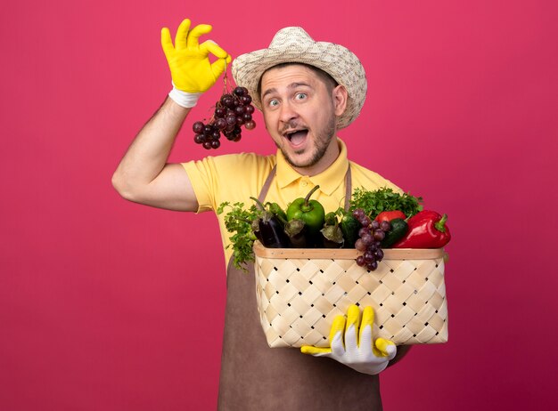 Young gardener wearing jumpsuit and hat in working gloves holding crate full of vegetables showing bunch of grape looking at front surprised standing over pink wall