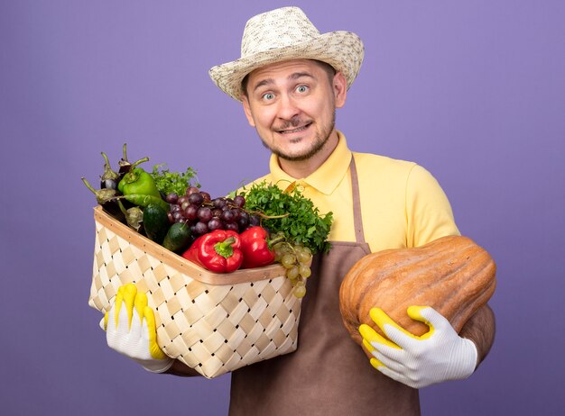 young gardener wearing jumpsuit and hat in working gloves holding crate full of vegetables with pumpkin looking at front smiling with happy face standing over purple wall