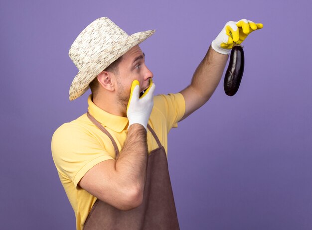 Young gardener wearing jumpsuit and hat in working gloves holding eggplant looking at it amazed and surprised standing over purple wall