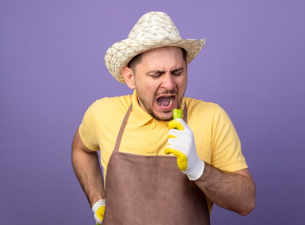 Young gardener wearing jumpsuit and hat in working gloves holding green chili pepper going to bite it standing over purple wall