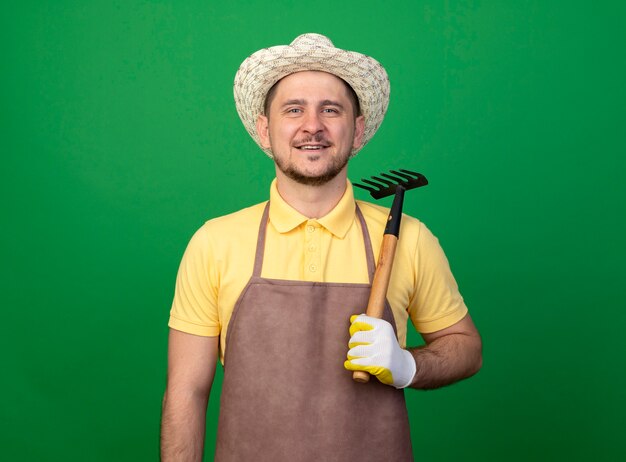 Young gardener wearing jumpsuit and hat in working gloves holding mini rake looking at front with smile on face standing over green wall